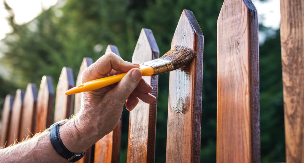 Close-up hand with paintbrush painting wooden picket fence outdoors